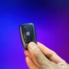 Compact remote control device held in hand against a vibrant purple background.