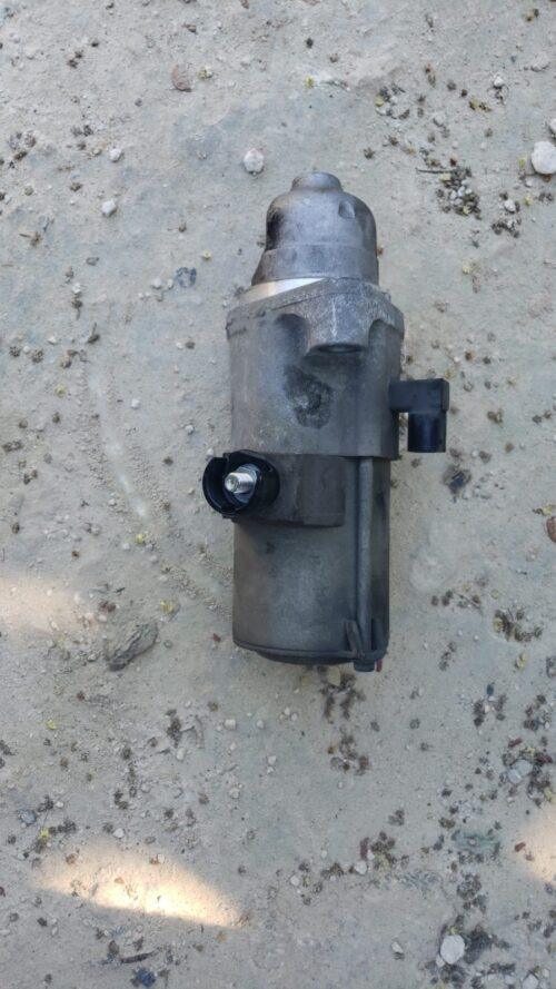 Close-up of a used automotive fuel pump lying on sandy ground, showing signs of wear and dirt.